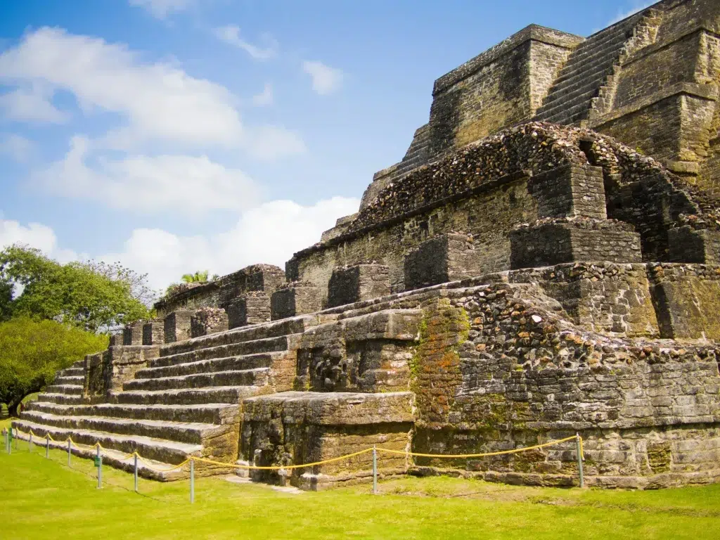 Ancient stone stepped pyramid ruins with broad stairs, mossy walls, and rope barriers on a grassy lawn under a blue sky.